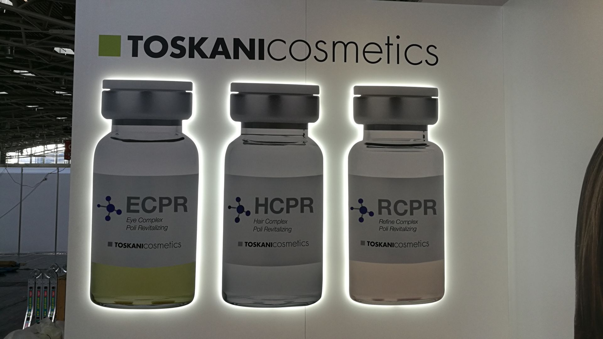 Schild für Toskani Cosmetics, drei Produktfläschchen, beleuchtet vor weißem Hintergrund.