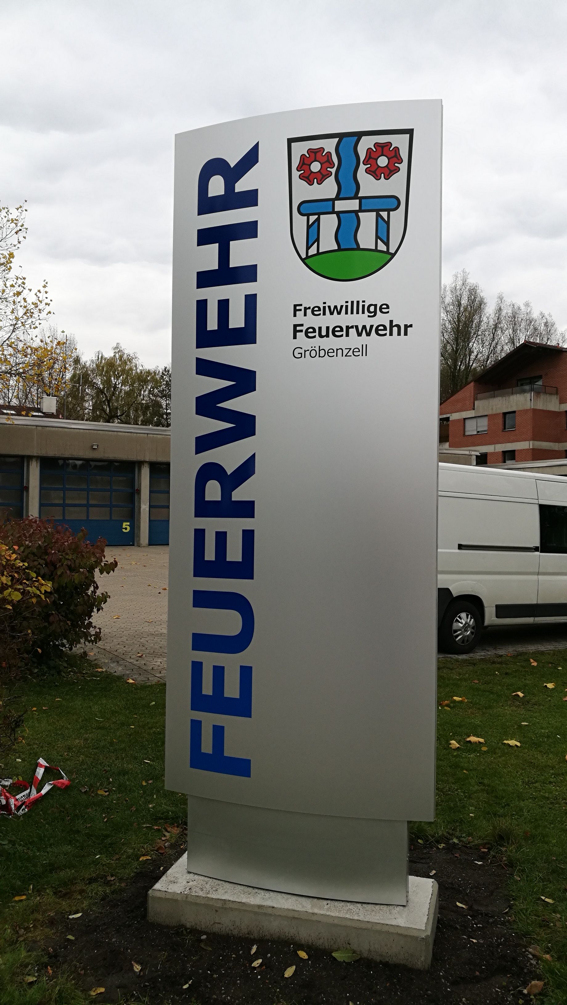 Schild „Feuerwehr“ mit blauer Schrift und Wappen, im Außenbereich.