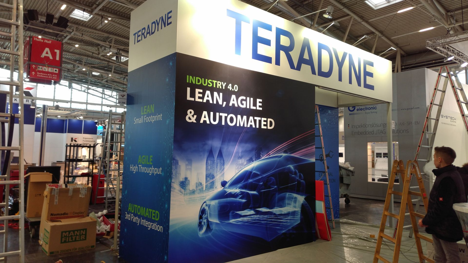 Messestand von Teradyne, Thema „Lean, Agile & Automated“, mit einer Autografik. Blau-, Weiß- und Orangetöne.