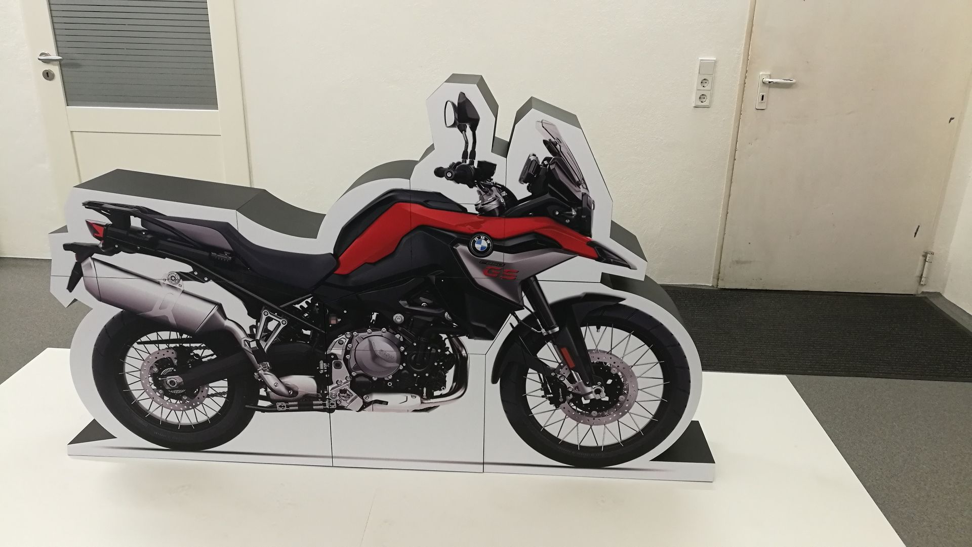 Pappaufsteller eines weiß-rot-schwarzen Motorrads auf einer weißen Fläche in einem Raum.