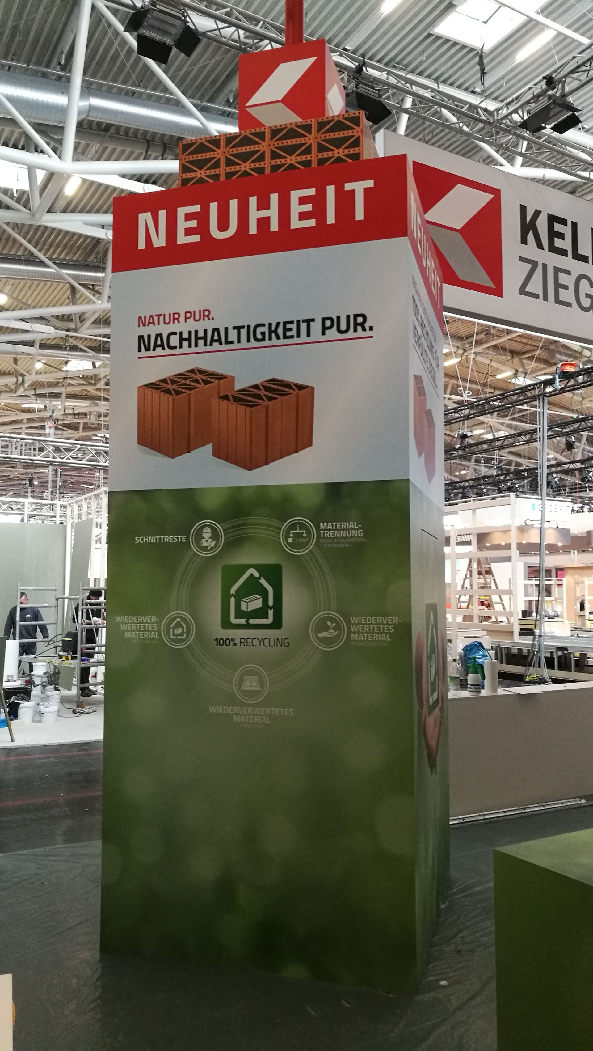 Ein hoher Werbeturm mit dem Wort „NEUHEIT“, der in einer Gebäudeumgebung für „Nachhaltigkeit pur“ wirbt.