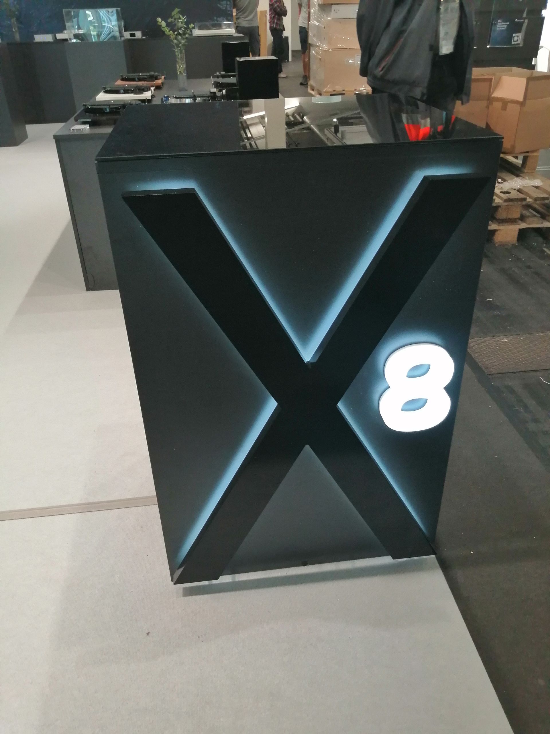 Schwarzer Kiosk mit beleuchtetem X und weißem „8“-Logo; grauer Boden.