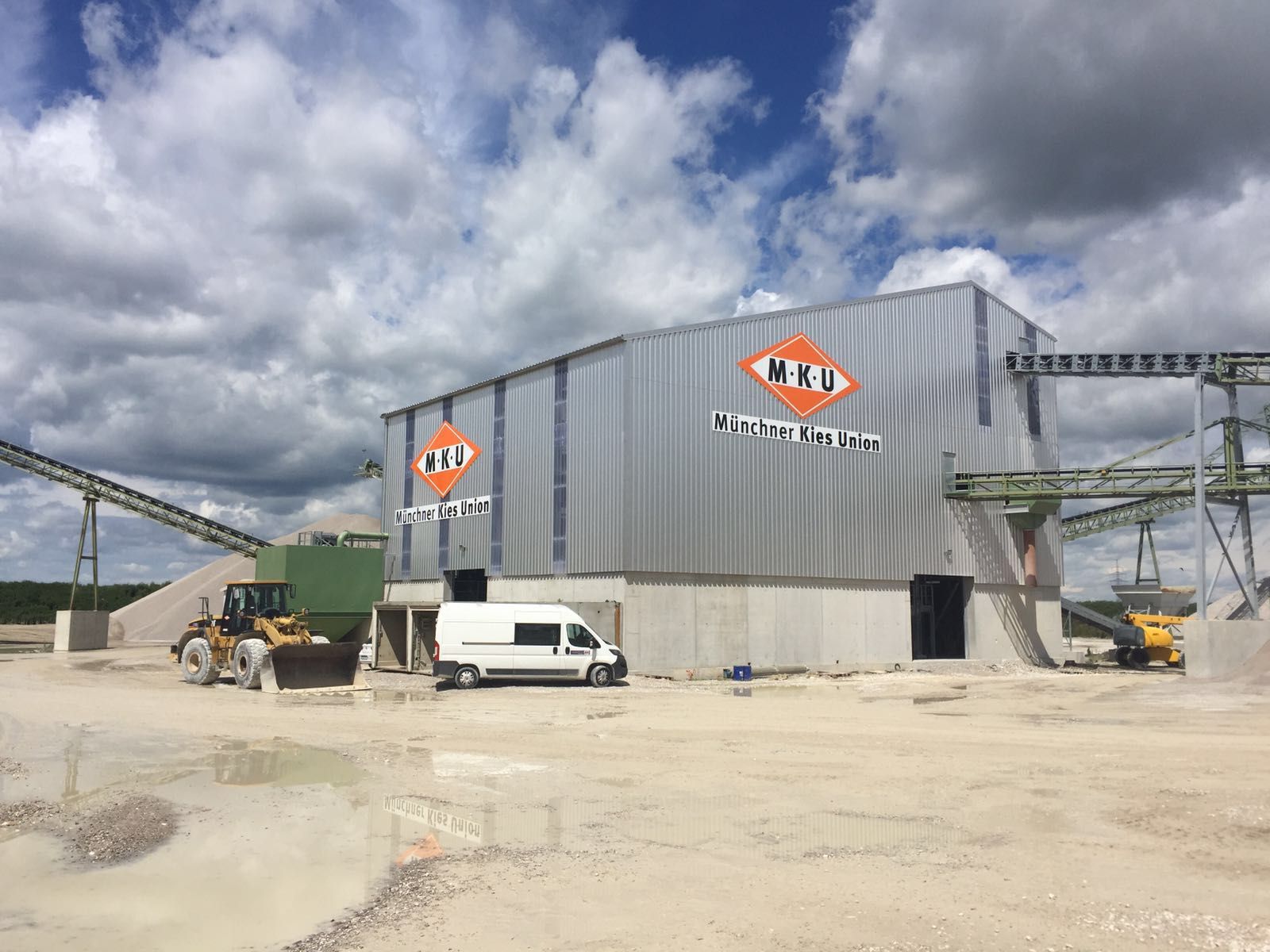 Fabrikgebäude mit Förderbändern und Baufahrzeugen, blauer Himmel, Logo.