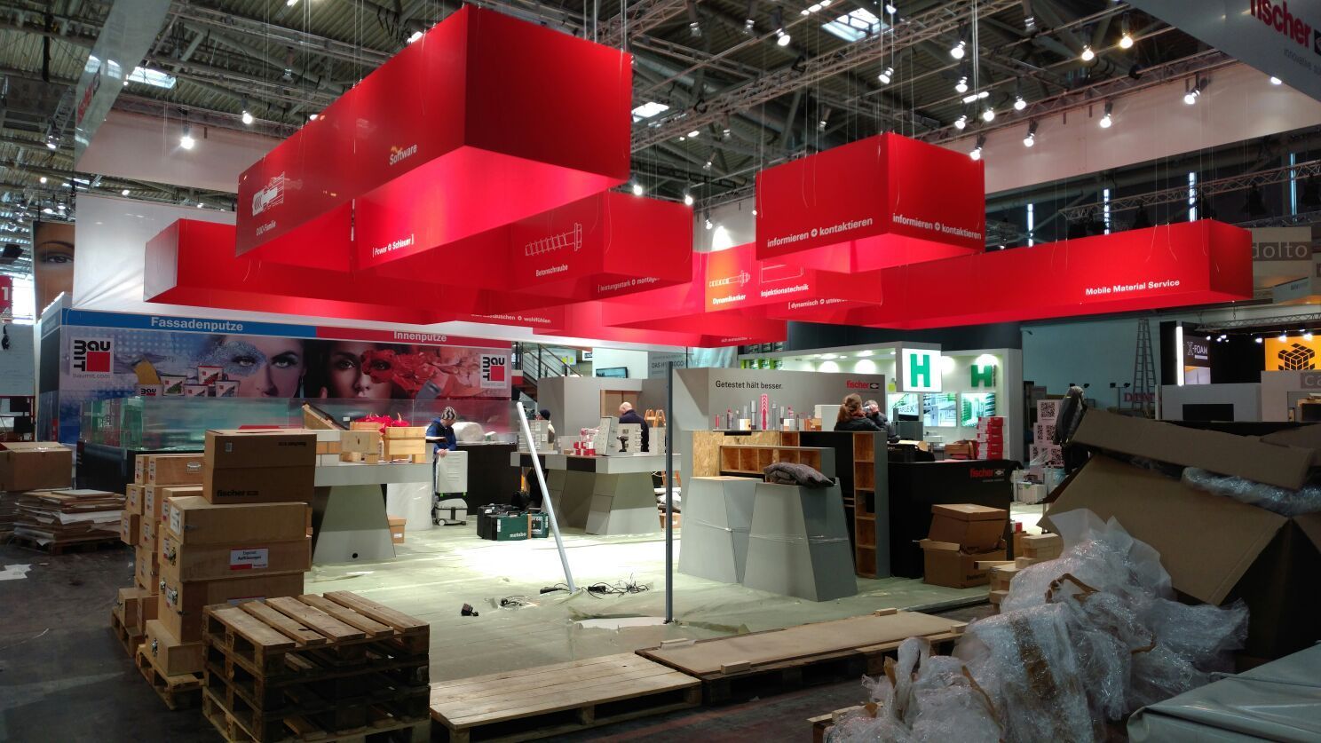 Rotes Deckendisplay an einem Messestand, im Vordergrund Kisten und Baumaterialien.