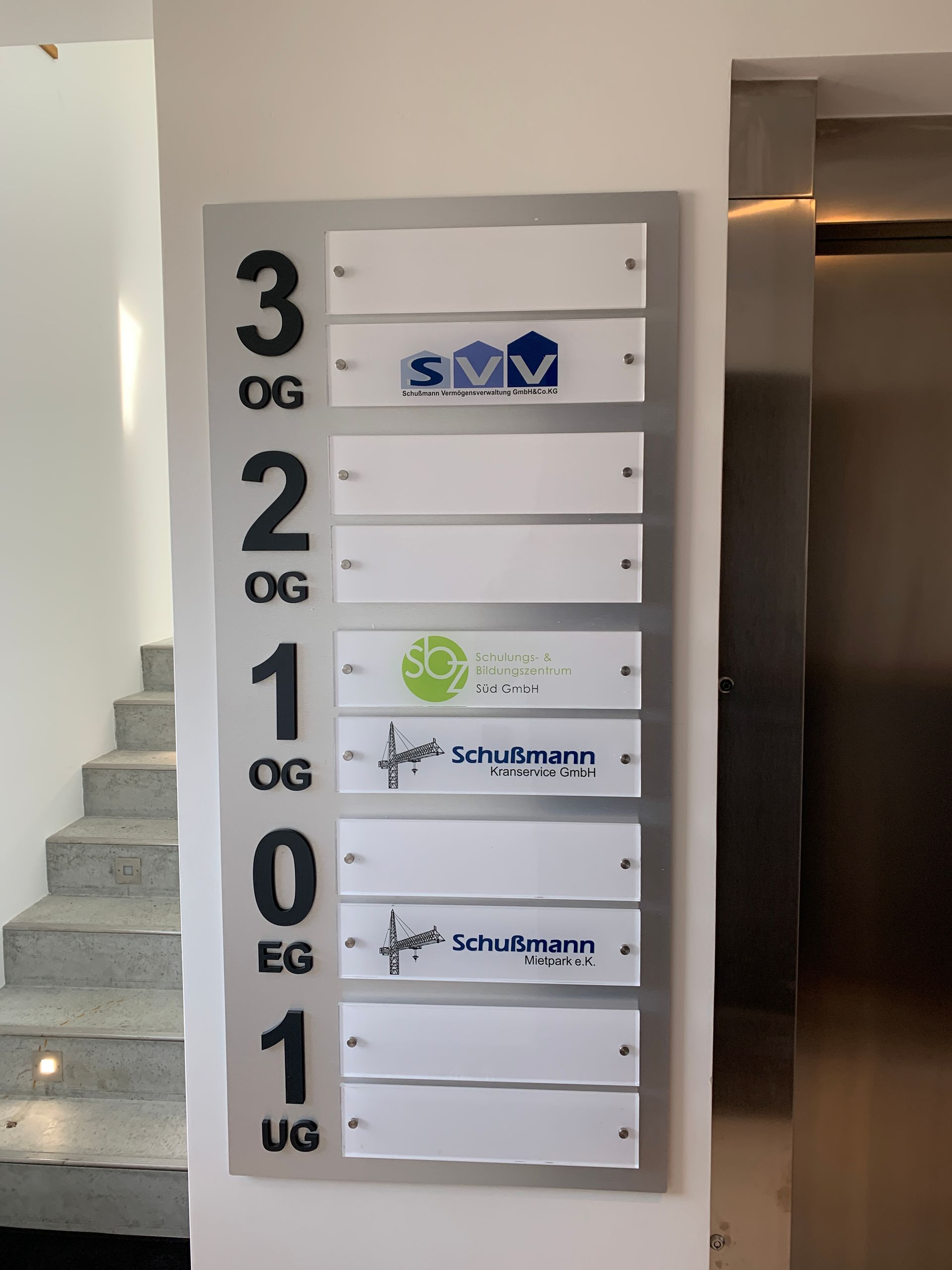 Wegweiser mit Firmenlogos und Stockwerken, neben einer Treppe und einem Aufzug.