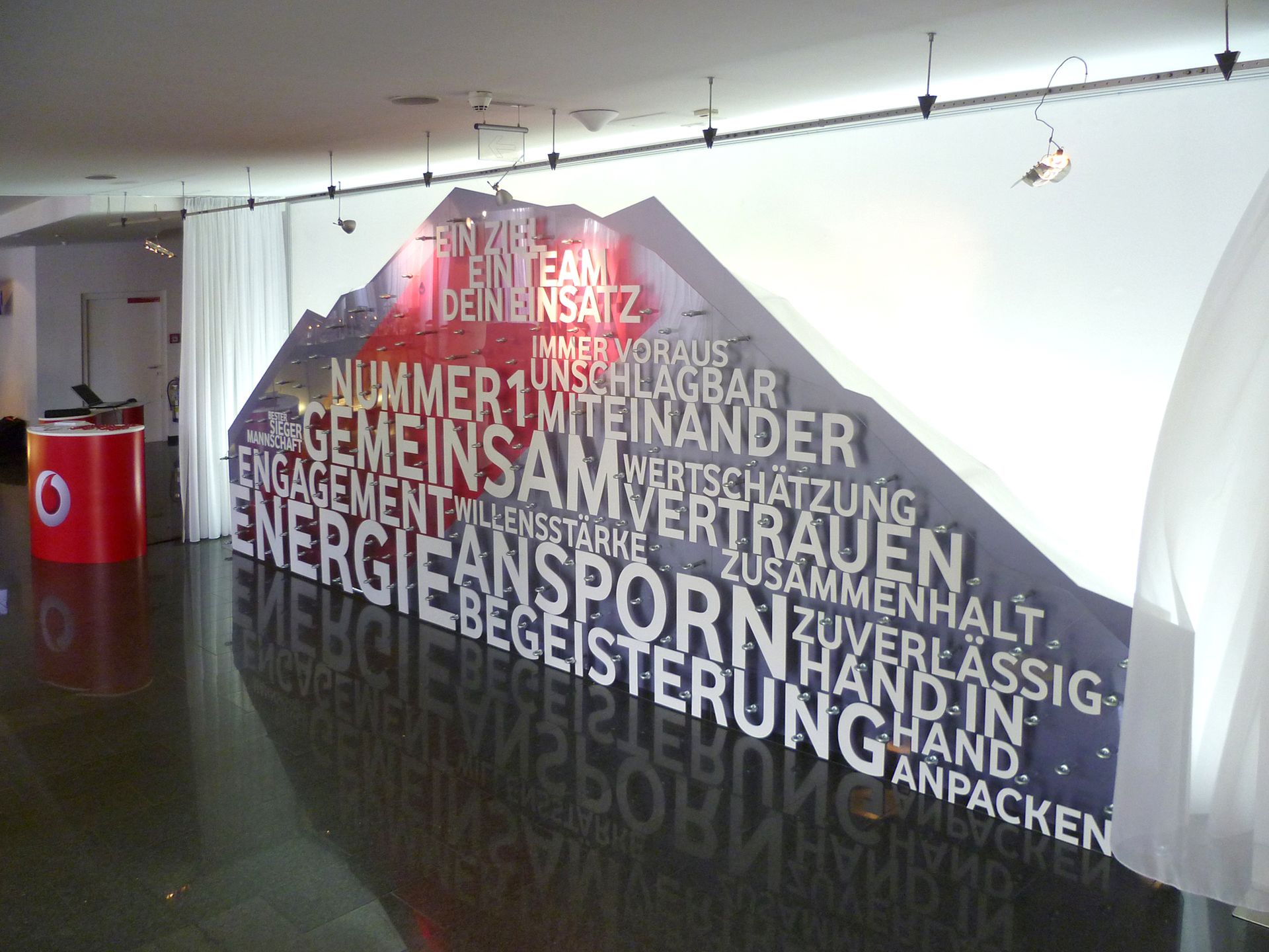 Großes Wandbild mit weißer 3D-Bergform, die deutsche Wörter wie „Gemeinsam“ und „Energie“ in einem Büro zeigt.