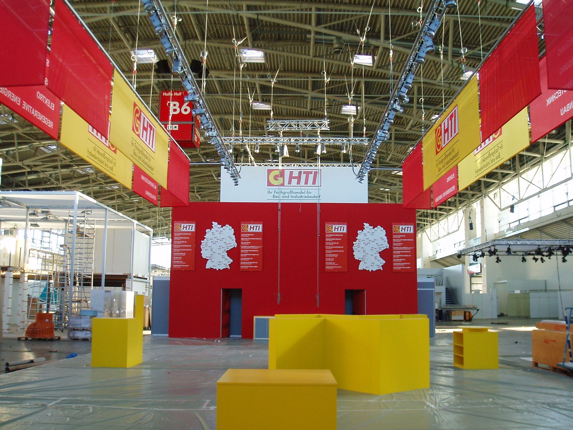 Große Halle mit rot-gelben HTT-Bannern, deutschen Kartendisplays, gelben Blöcken auf dem Boden.