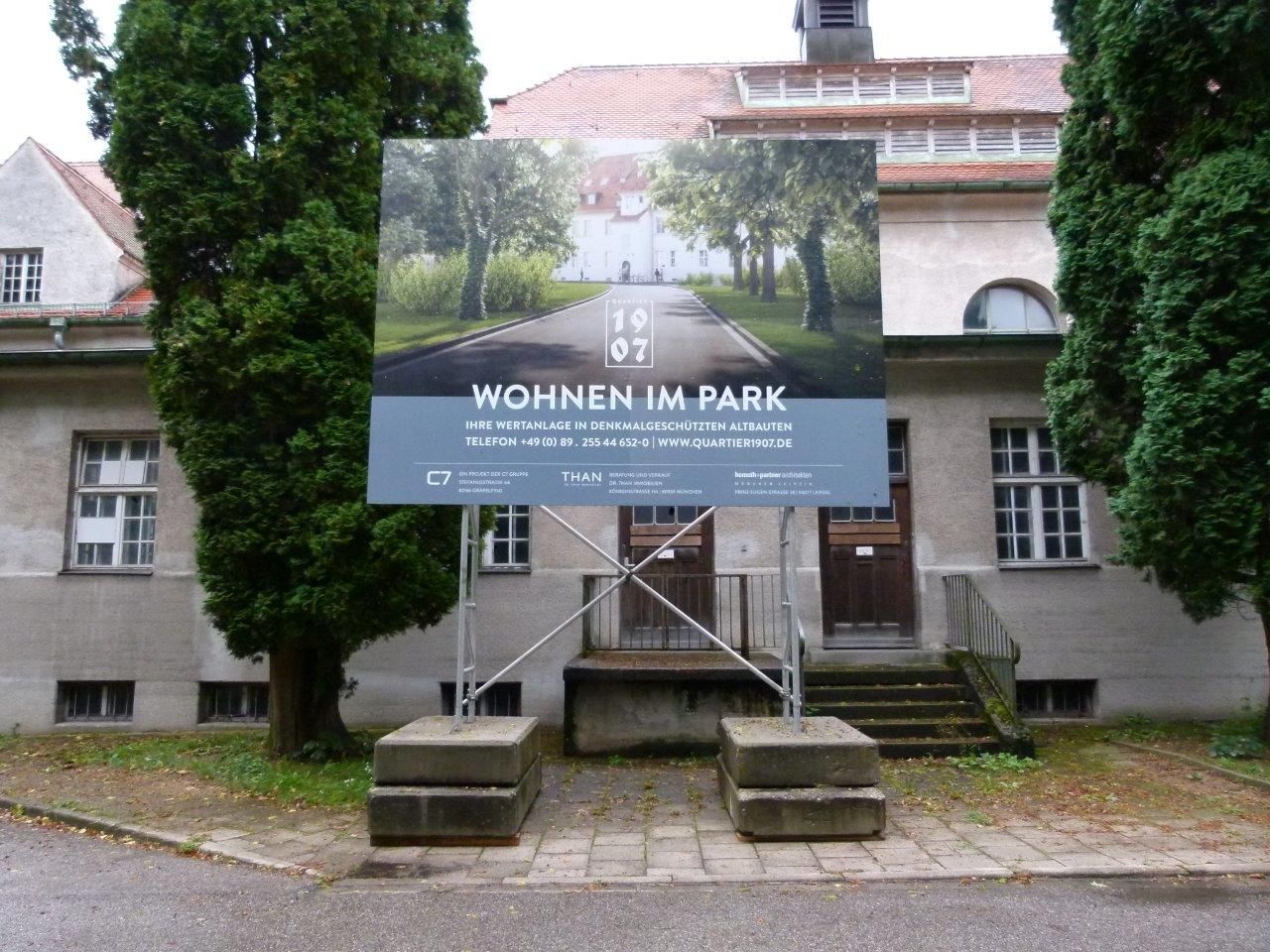 Schild mit der Aufschrift „Wohnen im Park“ vor einem historischen Gebäude mit Blick auf eine von Bäumen gesäumte Straße.