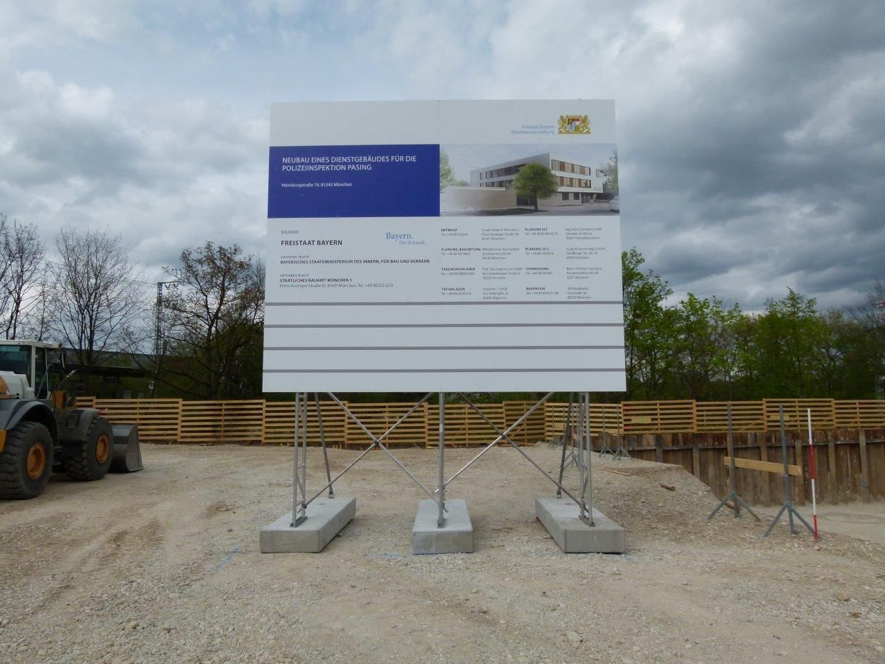 Baustellenschild mit Bauplänen, gestützt von Betonblöcken, Schotterboden, bewölkter Himmel.