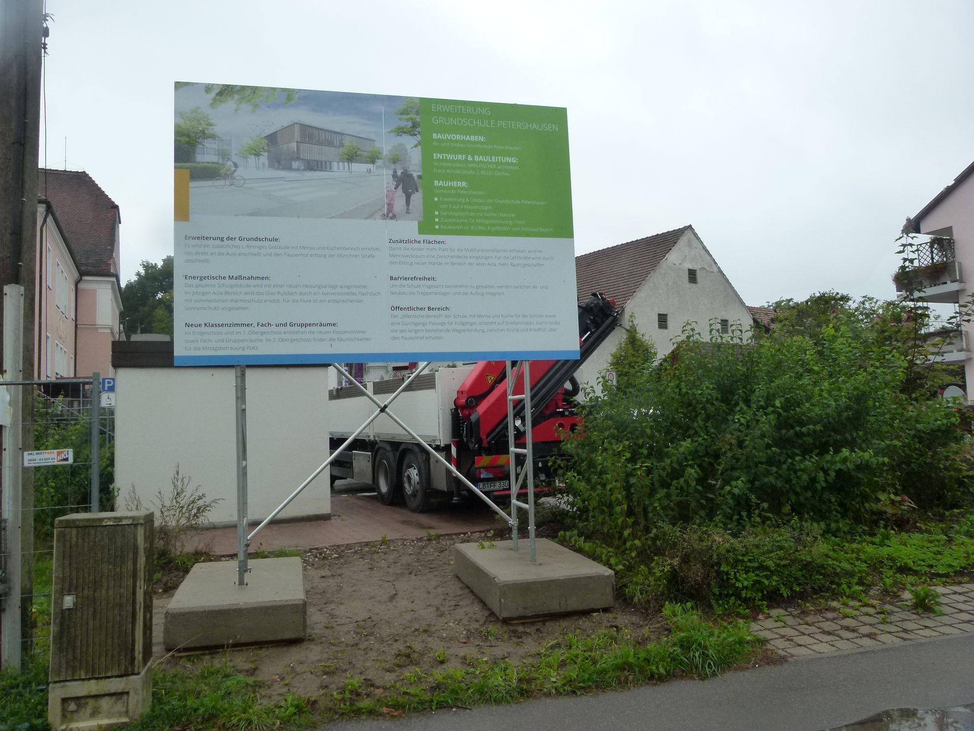 Bauschild in der Nähe eines Gebäudes, mit einem LKW im Hintergrund an einem bewölkten Tag.