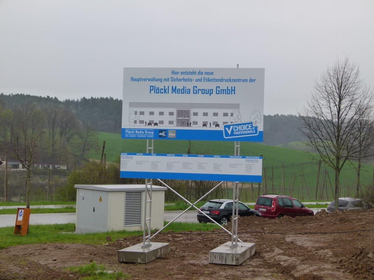 Werbetafel der Ploch Media Group GmbH vor einem Gebäude, Feld und Bäumen unter bewölktem Himmel.
