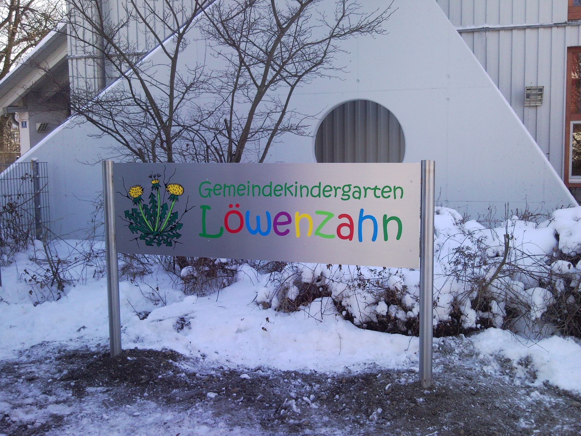 Sign for Gemeindekindergarten Löwenzahn (dandelion kindergarten) with colorful text and logo in front of snowy landscape.