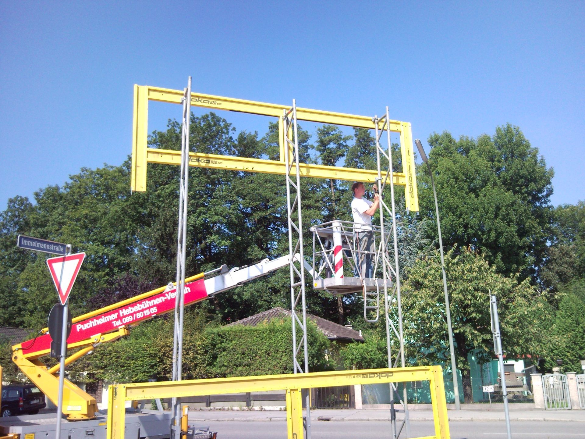 Ein Arbeiter installiert auf einer Hebebühne einen gelben Schilderrahmen über einer Straße.