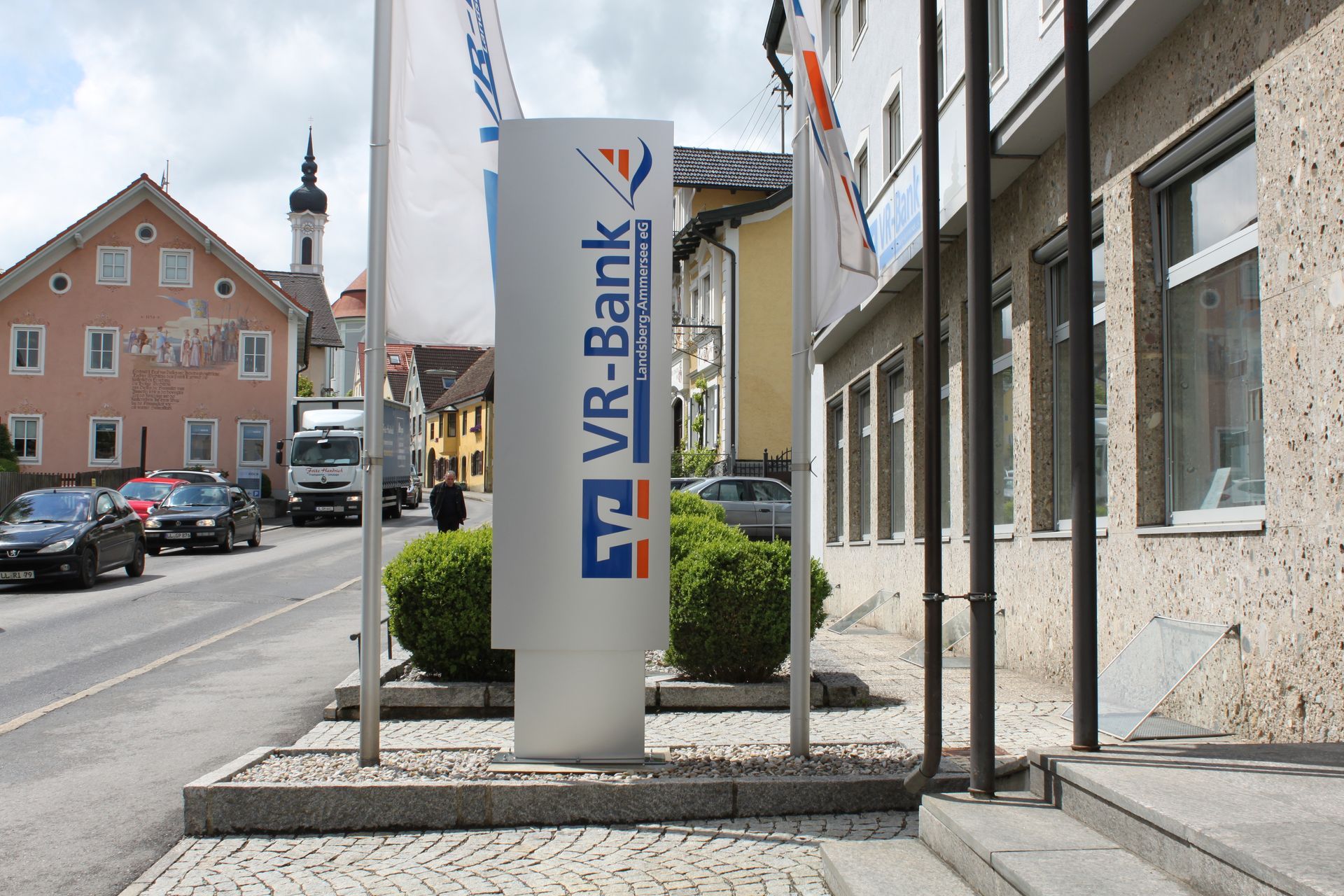 VR-Bank-Schild vor einem Gebäude an einer Straße; Autos, Flaggen und andere Gebäude im Hintergrund.