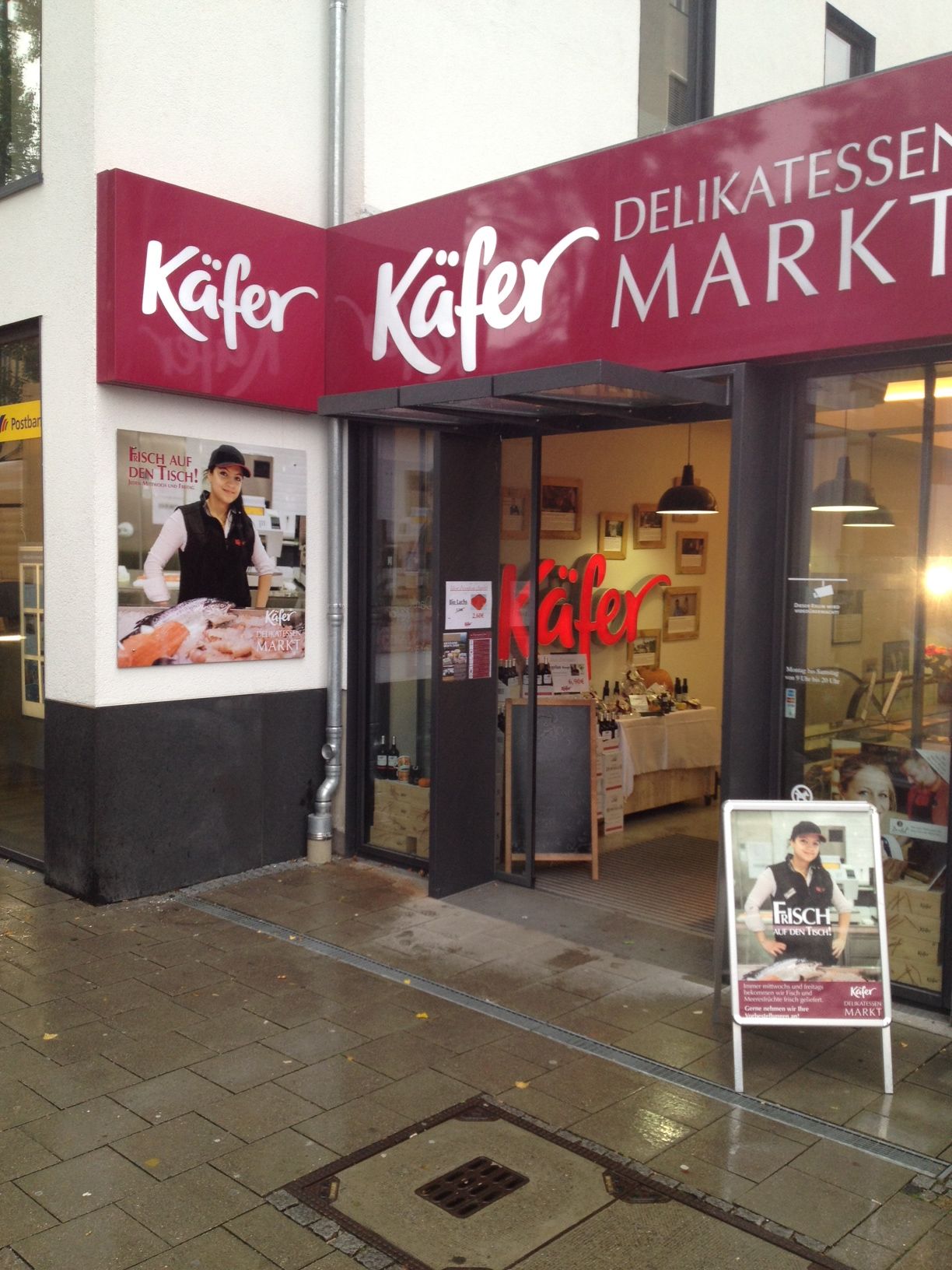 Eingang zum Käfer Delikatessen Markt. Rot-weißes Schild, Schaufenster mit Personenwerbung. Gepflasterter Bürgersteig.