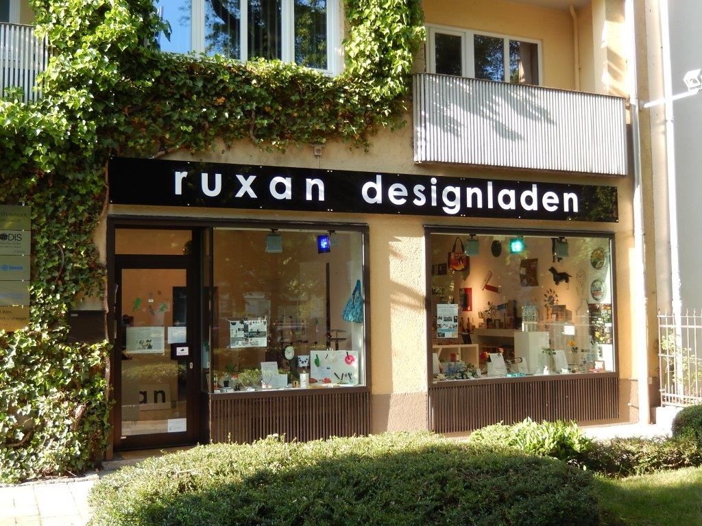 „Ruxan Designladen“-Schaufenster mit schwarzem Schild, Efeu und Schaufenstern, sonniger Tag.