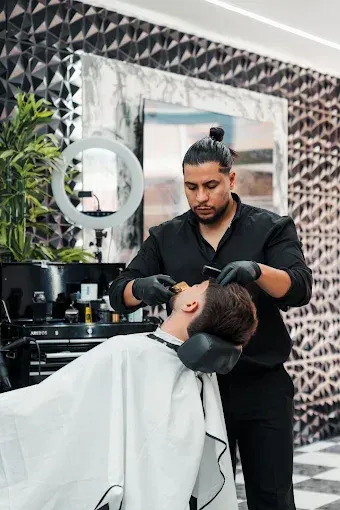 Un barbero afeitando la barba de un cliente en una barbería. Lleva guantes y ropa negros.