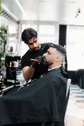 Un barbero le recorta la barba a un cliente en un salón. Ambos hombres visten de negro, uno con guantes. Interior luminoso.