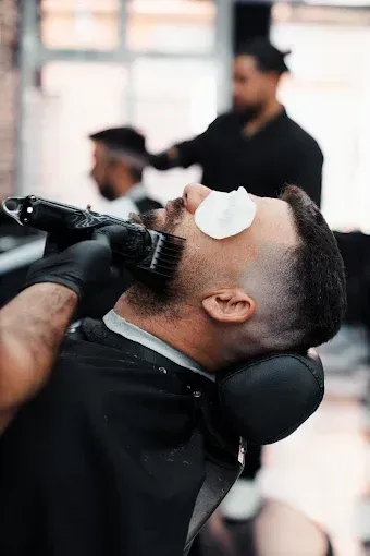 Un barbero recorta la barba de un cliente con una maquinilla en una barbería. Otro cliente al fondo.