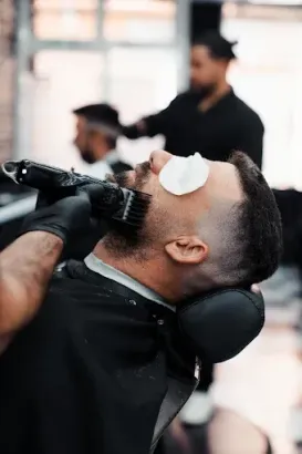Un barbero recorta la barba de un cliente con una maquinilla en una barbería. Otro cliente al fondo.