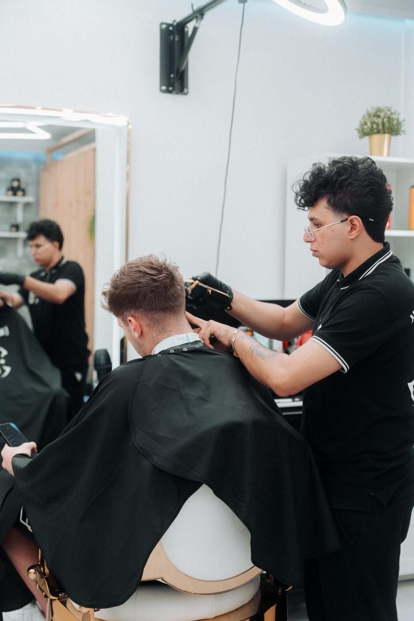 Un barbero corta el pelo a una clienta en un local bien iluminado. La clienta lleva una capa negra. 