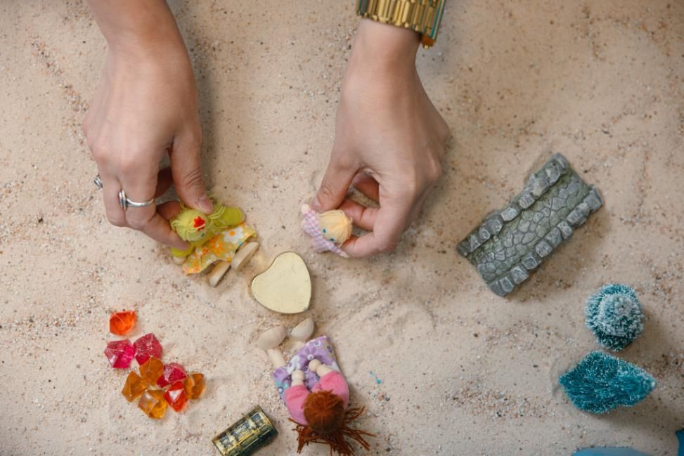 Manos organizando dulces de colores y pequeños objetos sobre la arena.