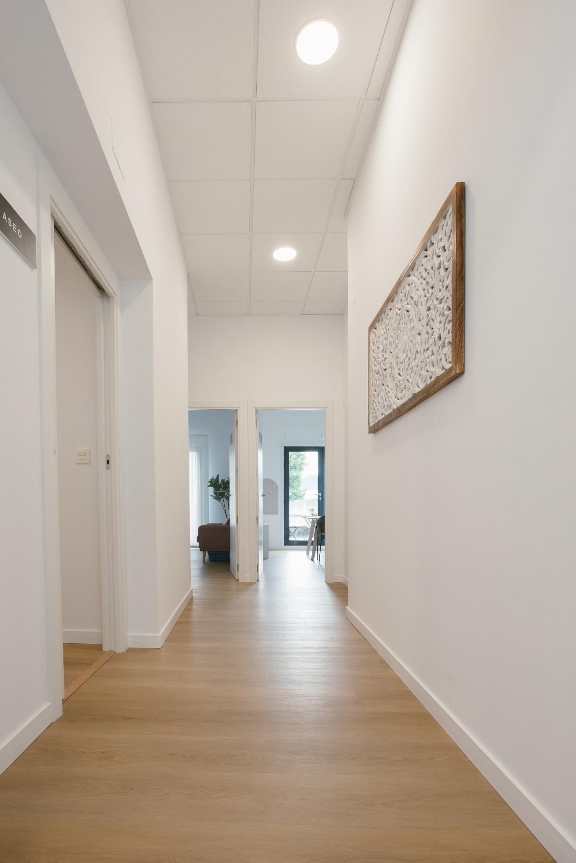 Niña sonriendo, jugando con muñecas en un taburete de madera con una persona con una camisa blanca en una habitación con ventana.