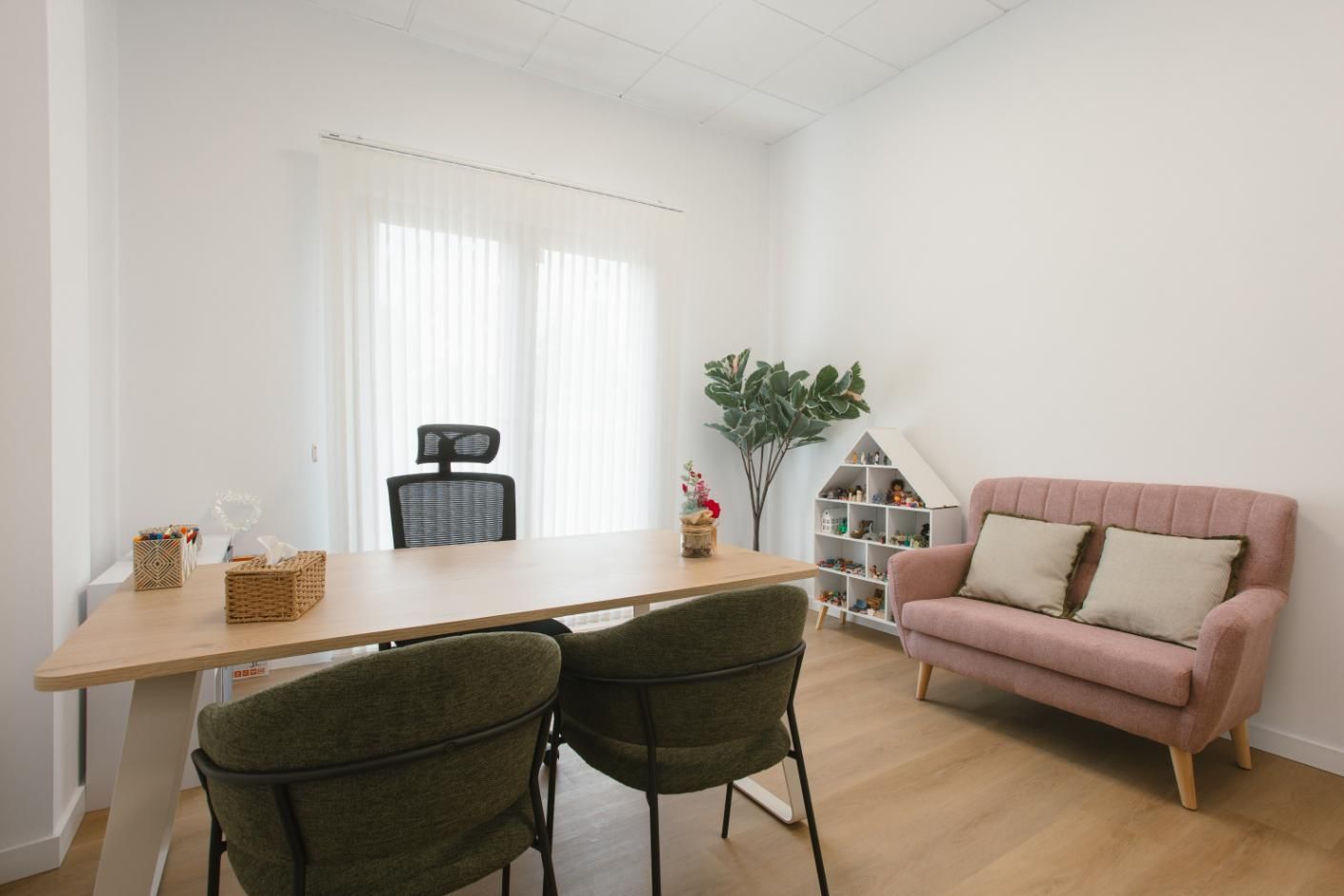 Consultorio del terapeuta: escritorio con dos sillas, sofá rosa, casita de juegos y una ventana con persianas.