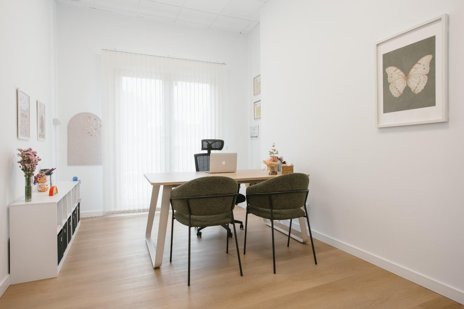 Niña sonriendo, jugando con muñecas en un taburete de madera con una persona con una camisa blanca en una habitación con ventana.