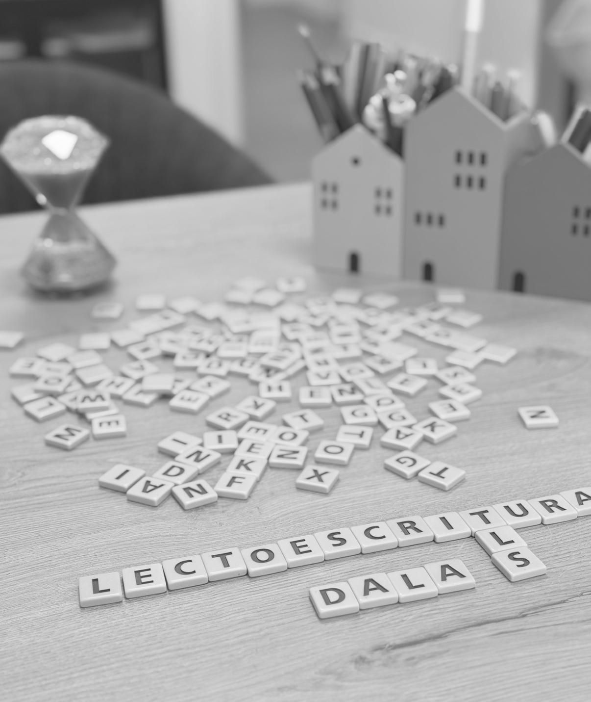 Niña sonriendo, jugando con muñecas en un taburete de madera con una persona con una camisa blanca en una habitación con ventana.