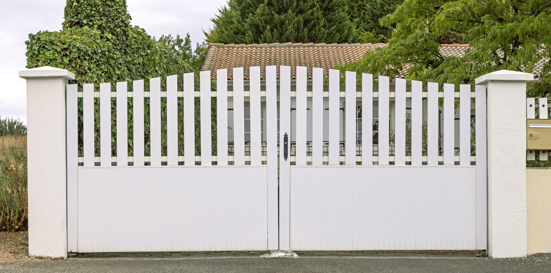 Portail d'allée en métal blanc avec barres verticales et piliers blancs devant une maison et des arbres