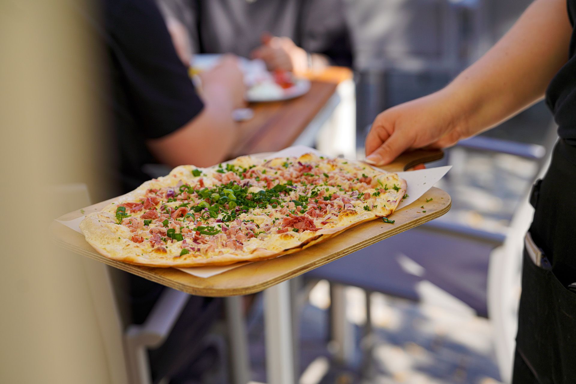 Flammkuchen auf einem Holzbrett wird vom Servicepersonal zu Gästen im Außenbereich getragen