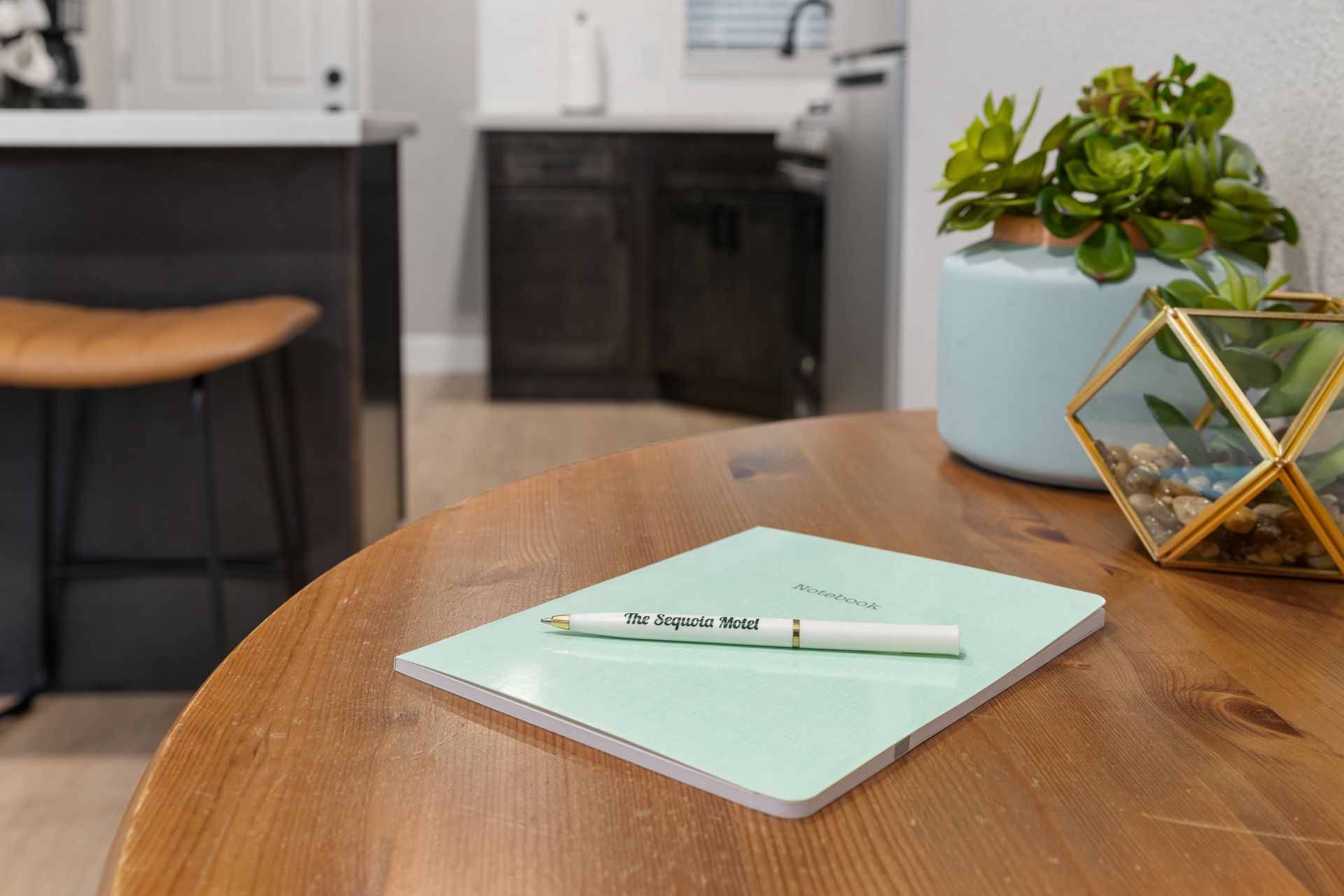 A light green notebook and pen on a wooden table, with a kitchen background and plants.