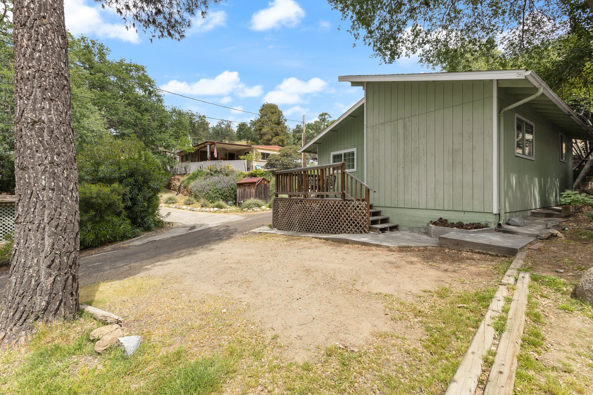 Green cabin with a gravel driveway and wooden deck in a wooded area.