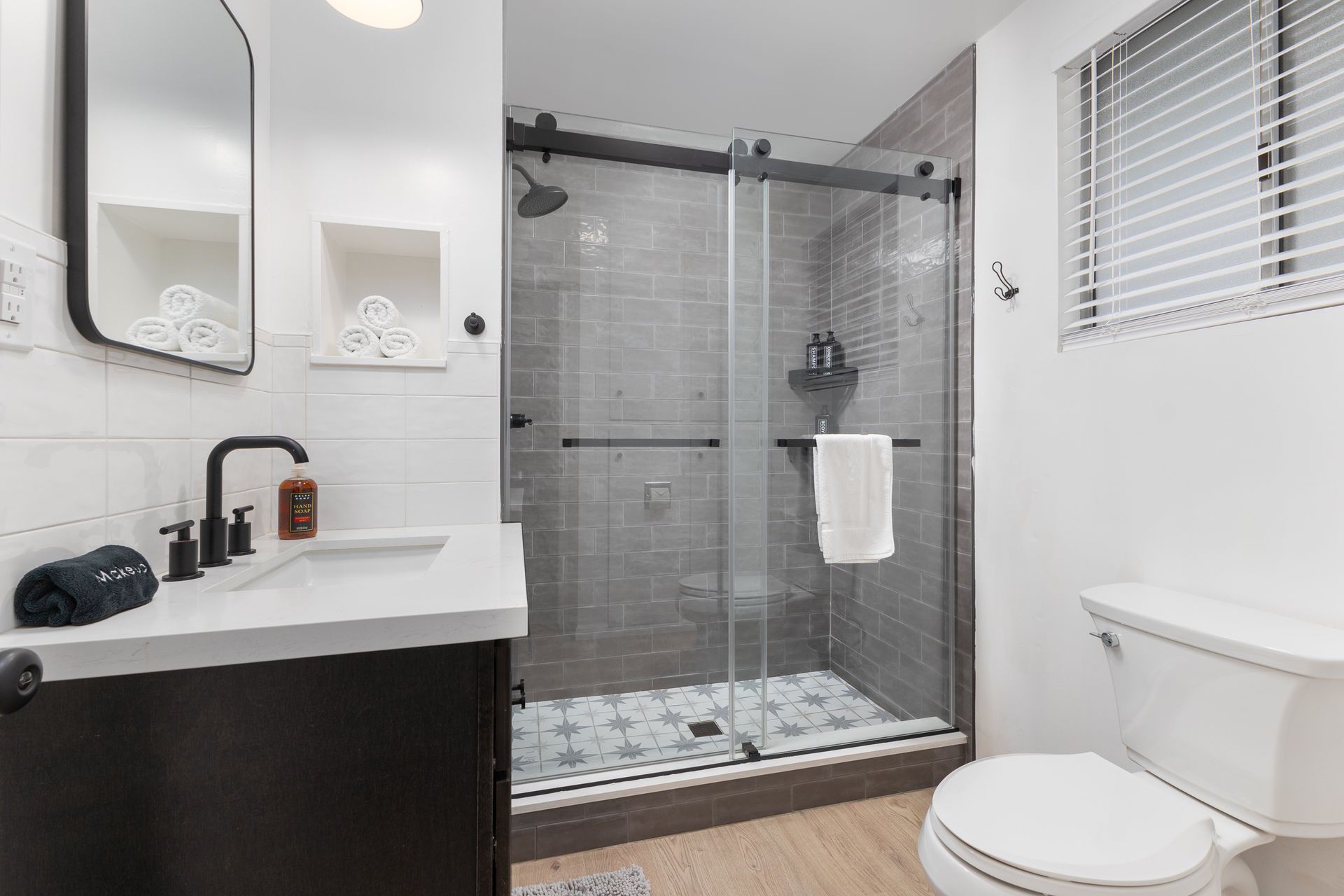 Modern bathroom: shower with gray tile, black hardware, white sink, dark cabinet, toilet, and window.