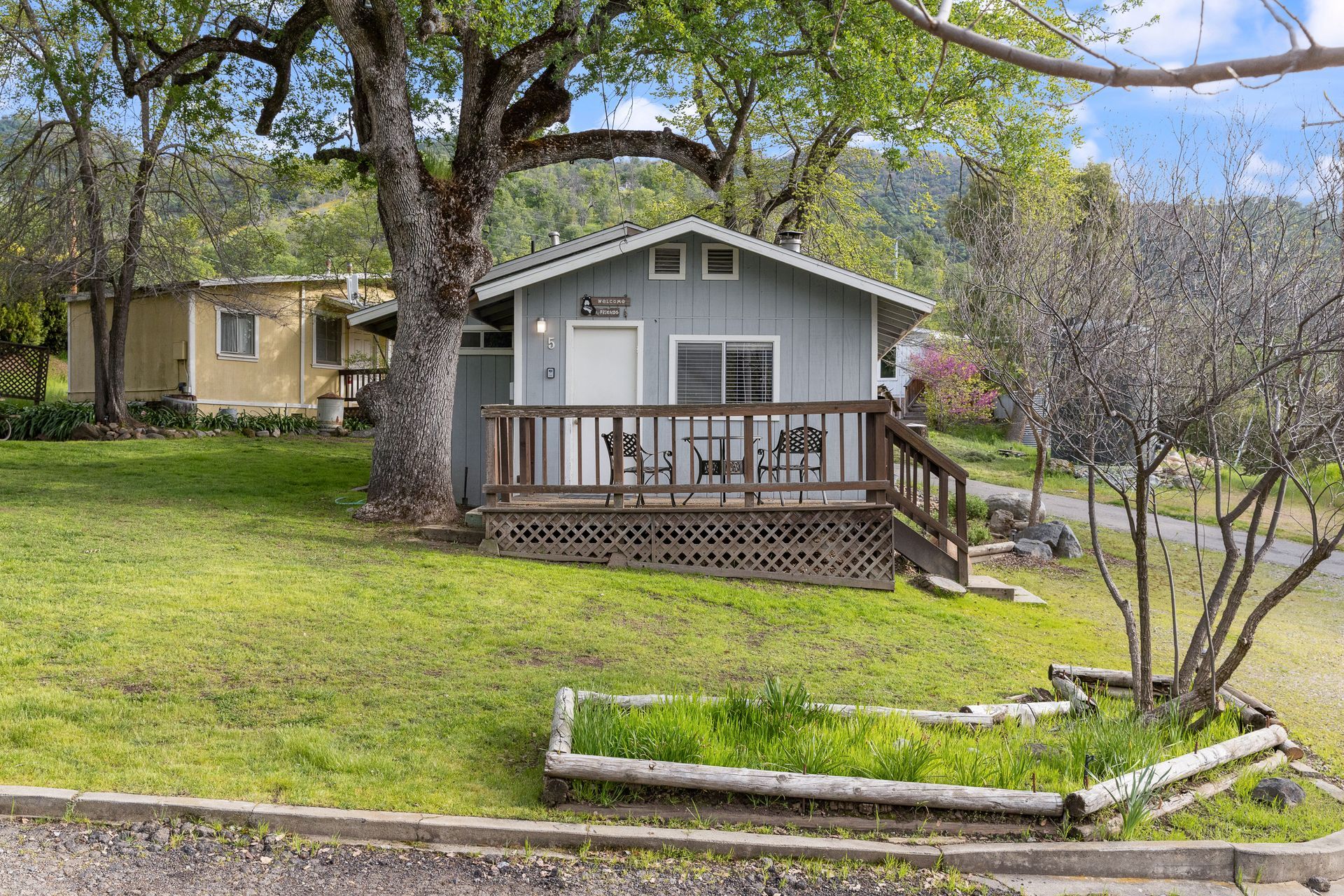 Small blue house with a wooden deck and a raised garden, in front of trees and a yellow building.