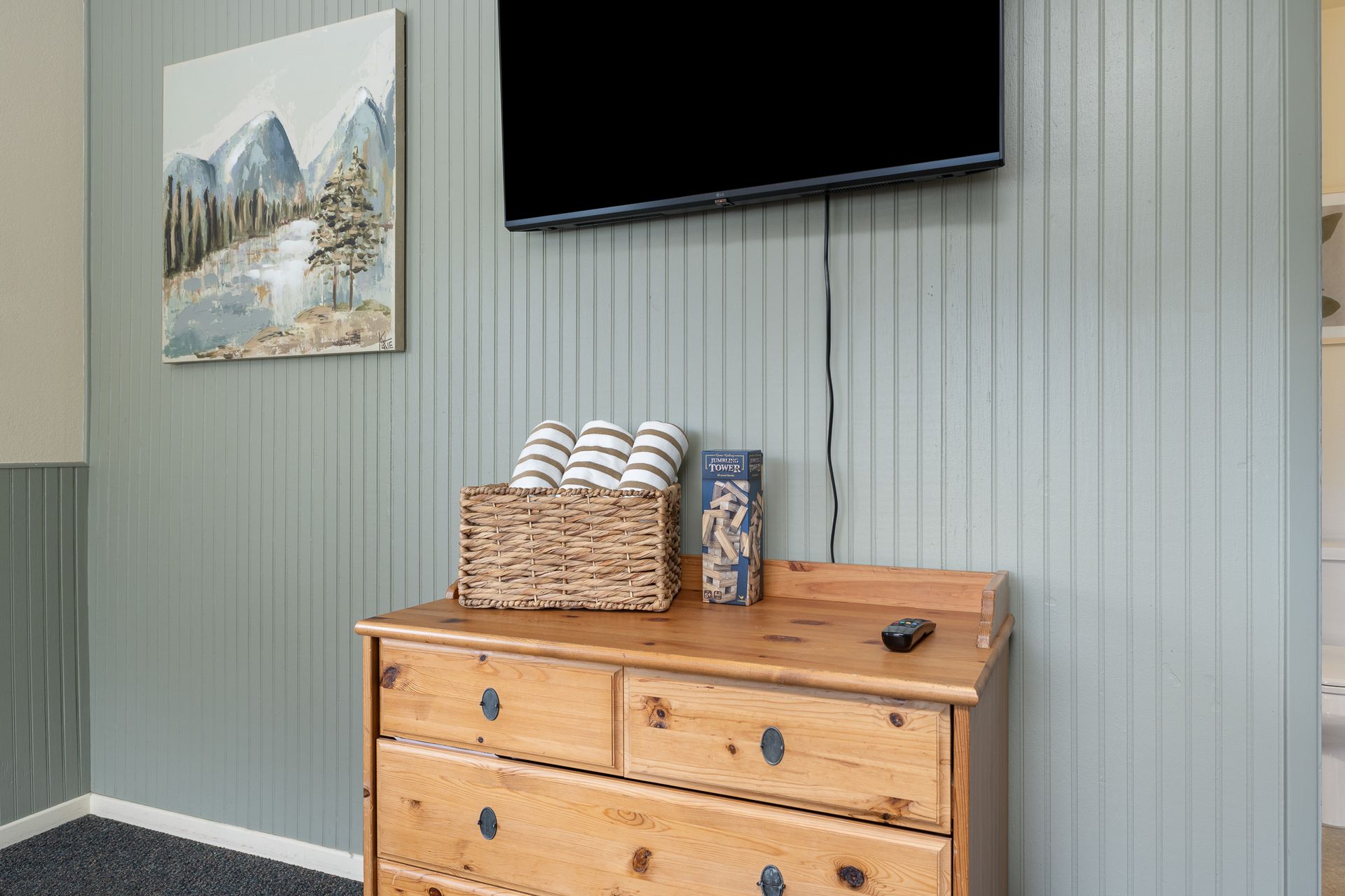 Wooden dresser with basket of towels and mounted TV on a light green paneled wall.