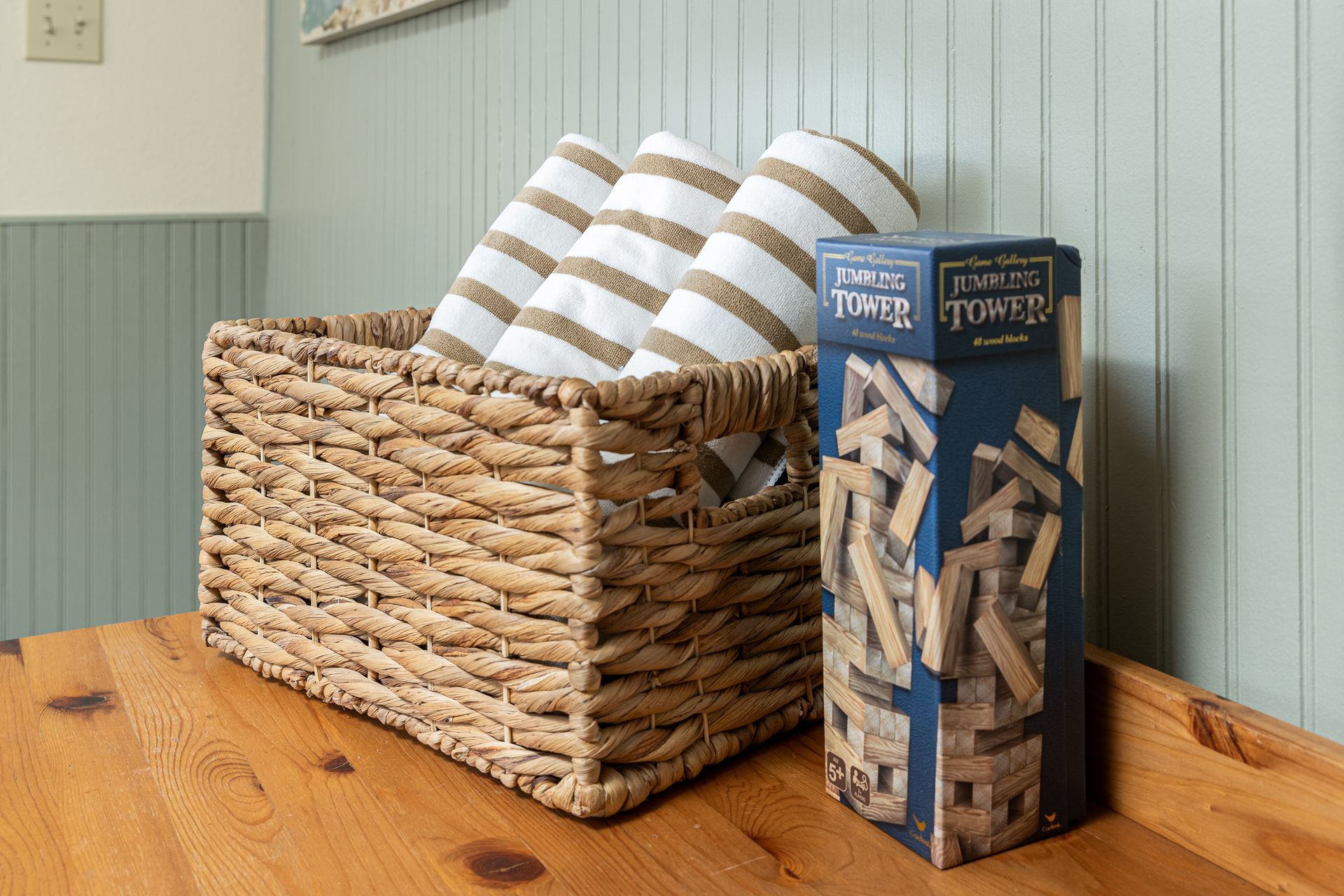 Woven basket with rolled towels next to a Jenga game on a wooden surface, against a blue wall.