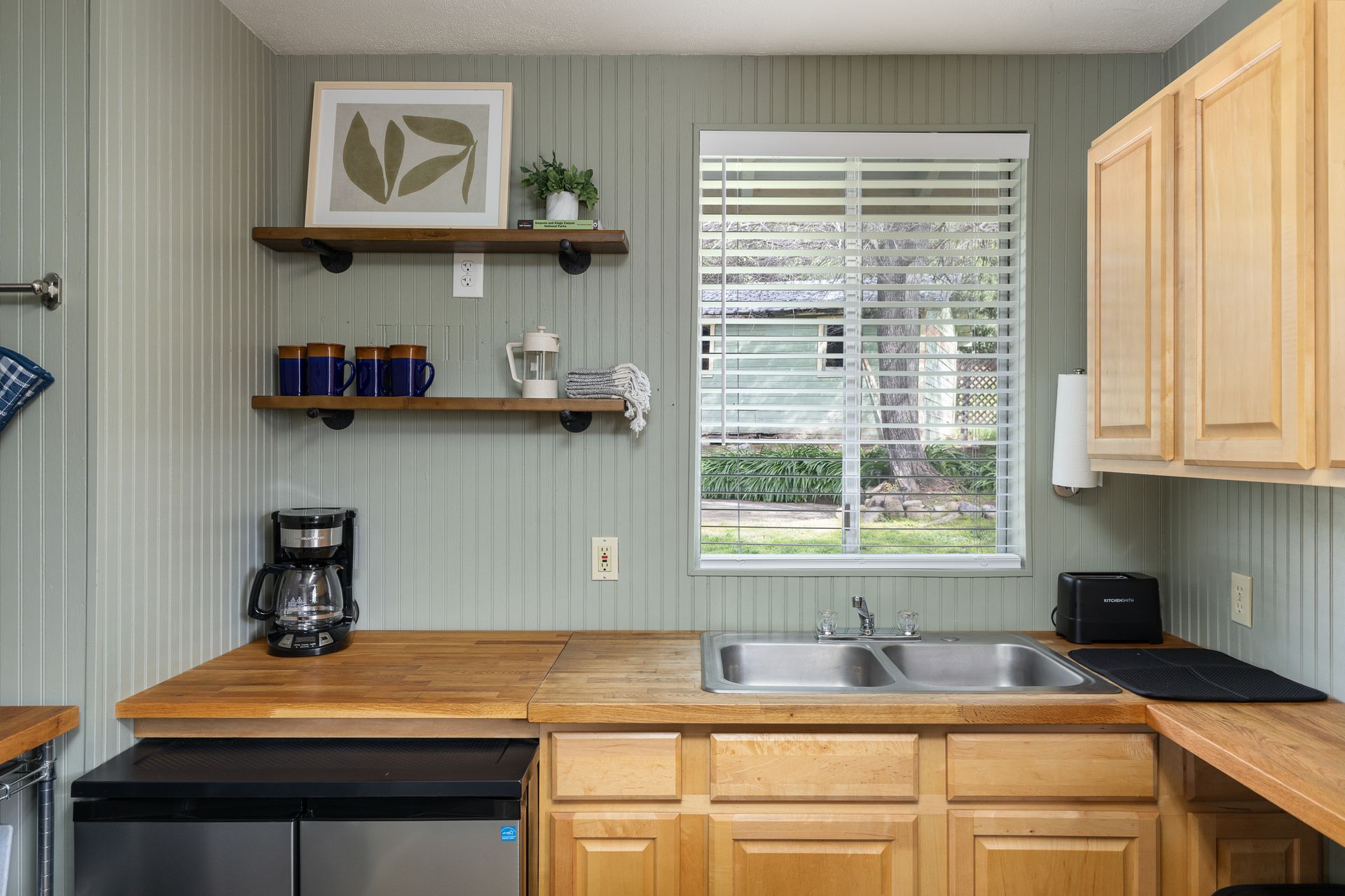 Small kitchen with light wood cabinets, sage green paneled walls, and open shelving.