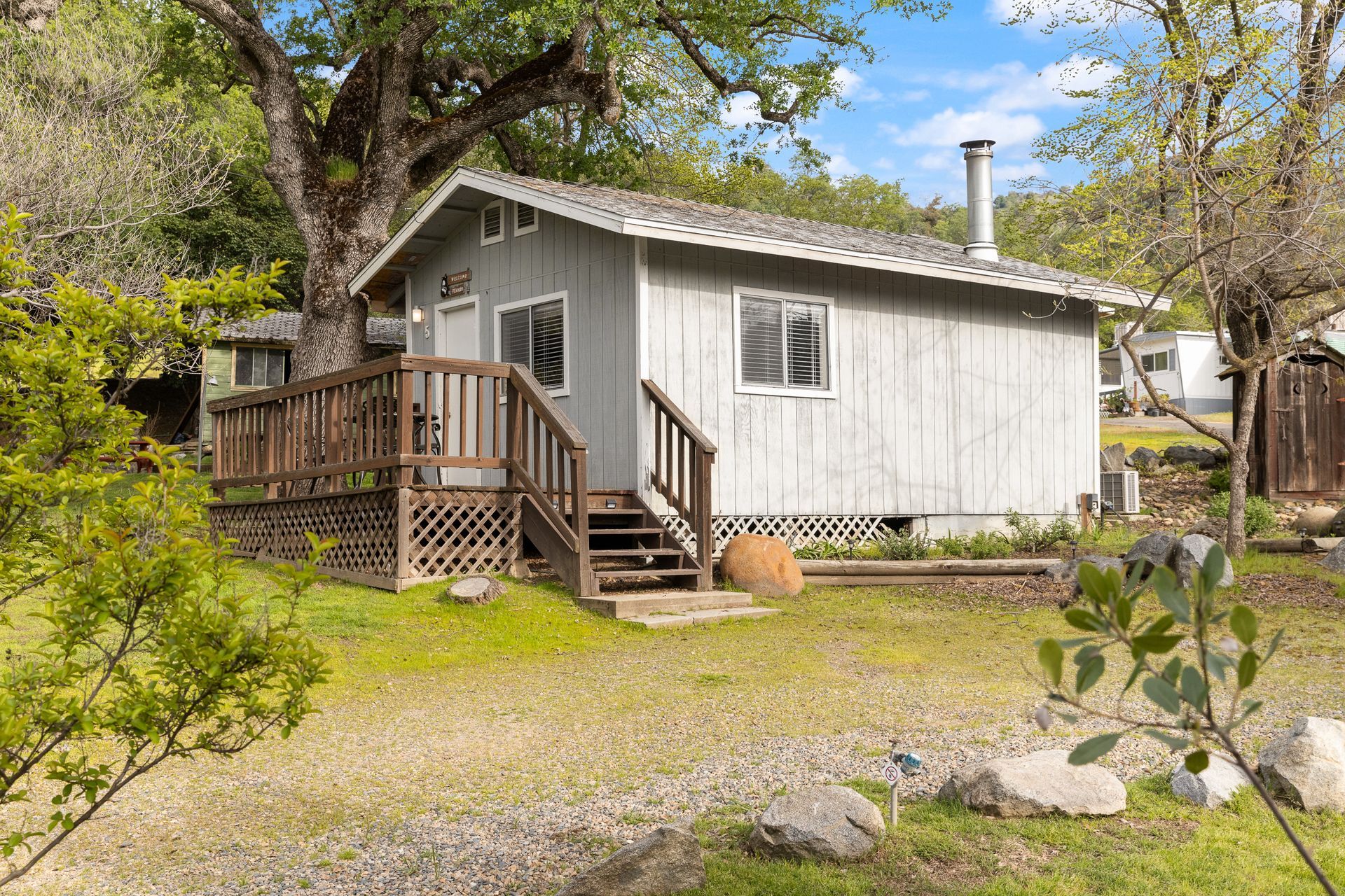 A small, gray cabin with a wooden deck, nestled amongst trees, with grassy yard.