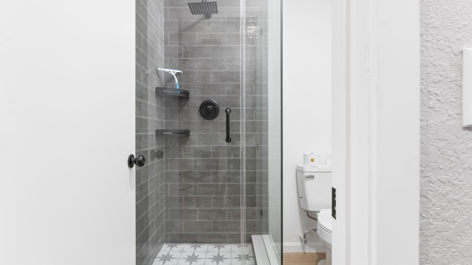 Gray-tiled shower with glass door and black fixtures; toilet in the small, neutral-toned bathroom.