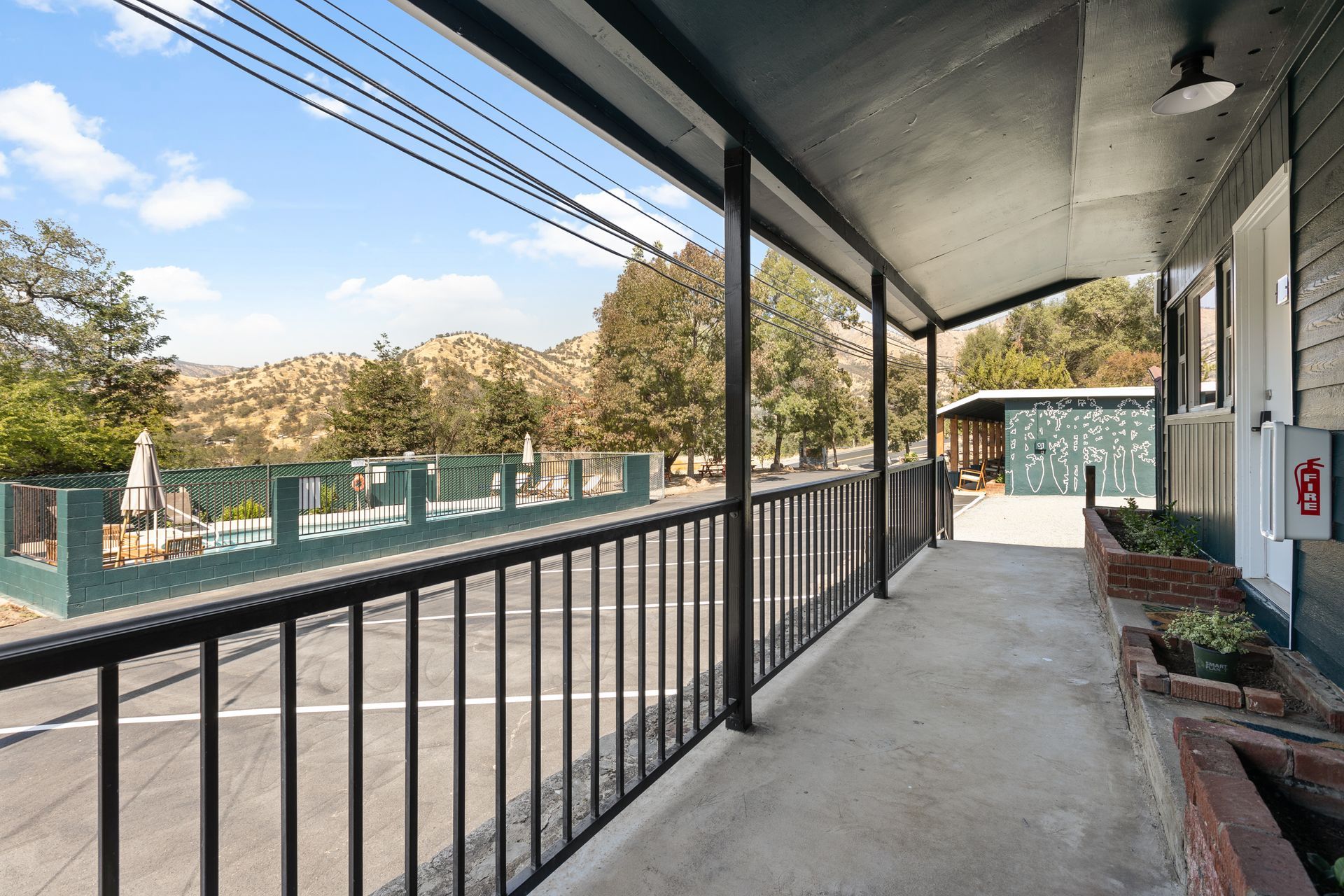 Long porch with black railing overlooking a street and a pool area, with mountains in the background.