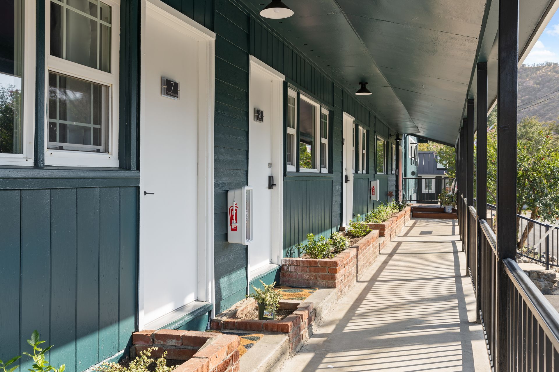 A row of teal motel rooms with white doors and a shaded walkway.