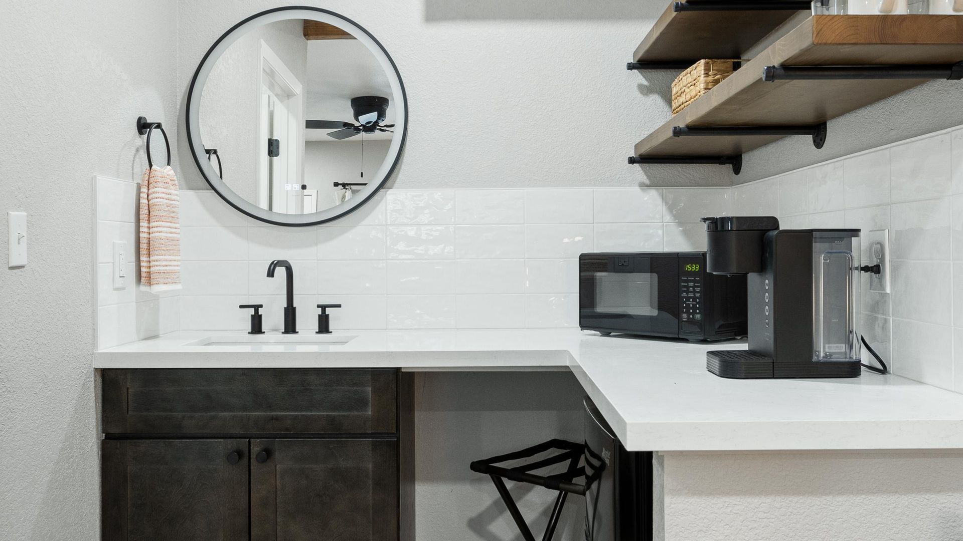 A small kitchen area with white countertops, black appliances, a round mirror, and dark cabinets.
