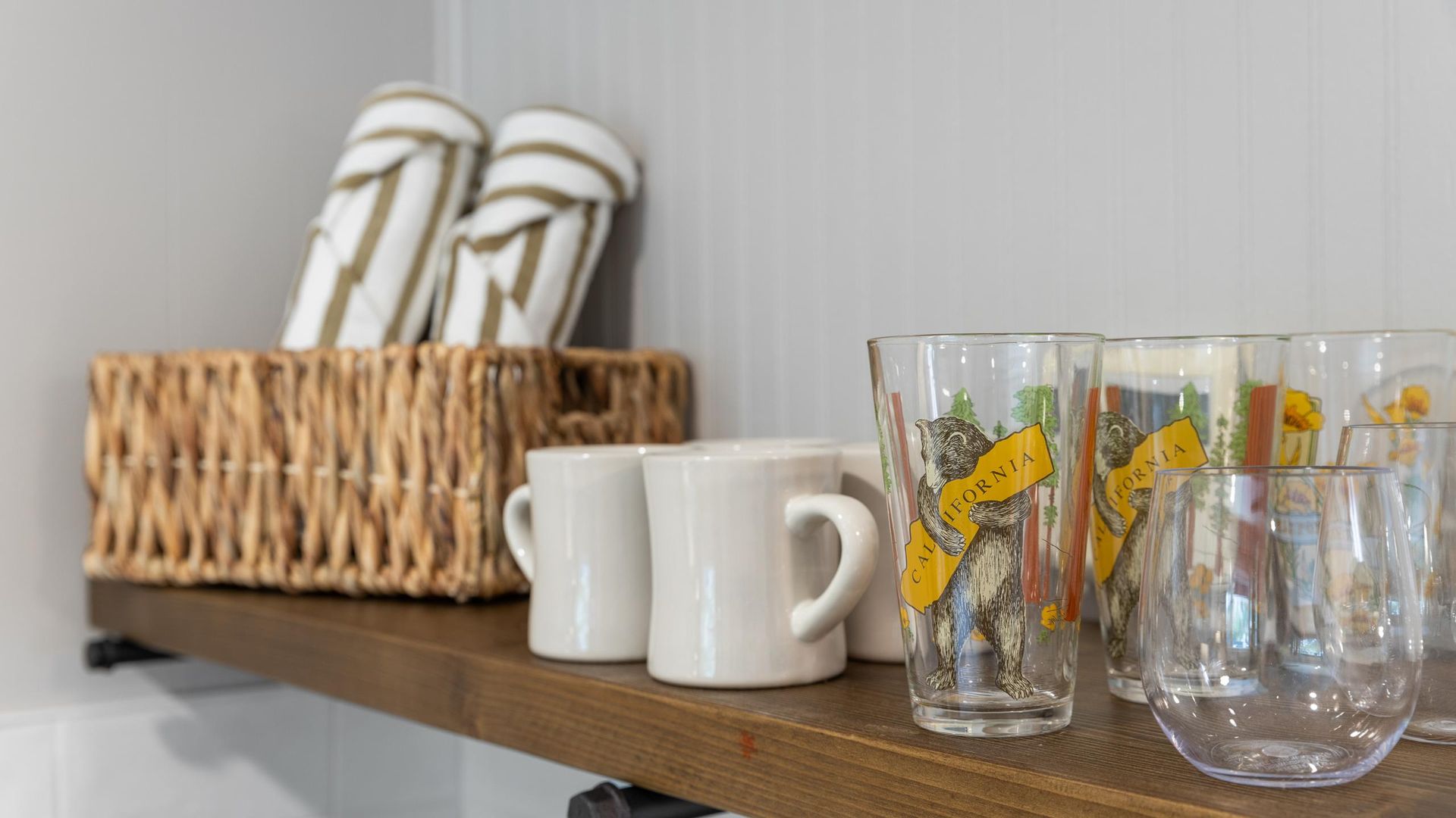 Shelf with rolled towels in basket, white mugs, printed and clear drinking glasses.