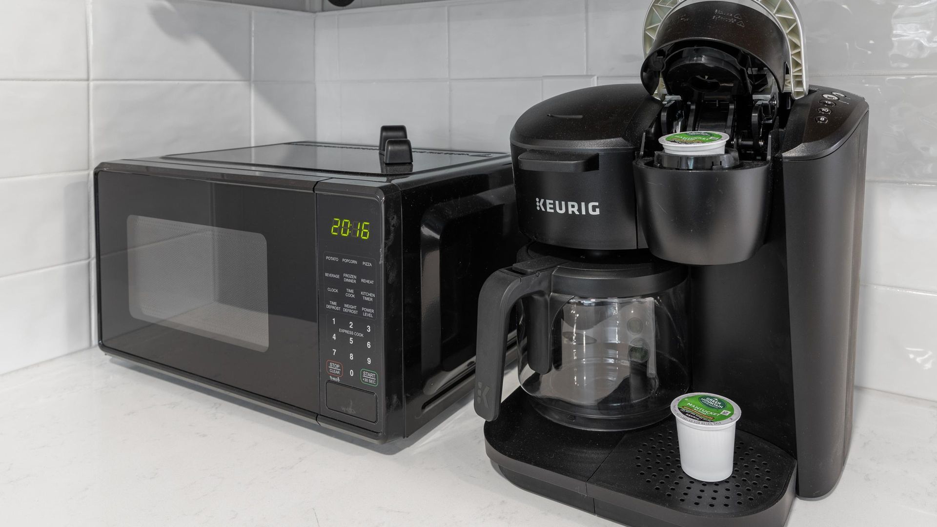 A black Keurig coffee maker and microwave sit on a white countertop next to each other in a kitchen.