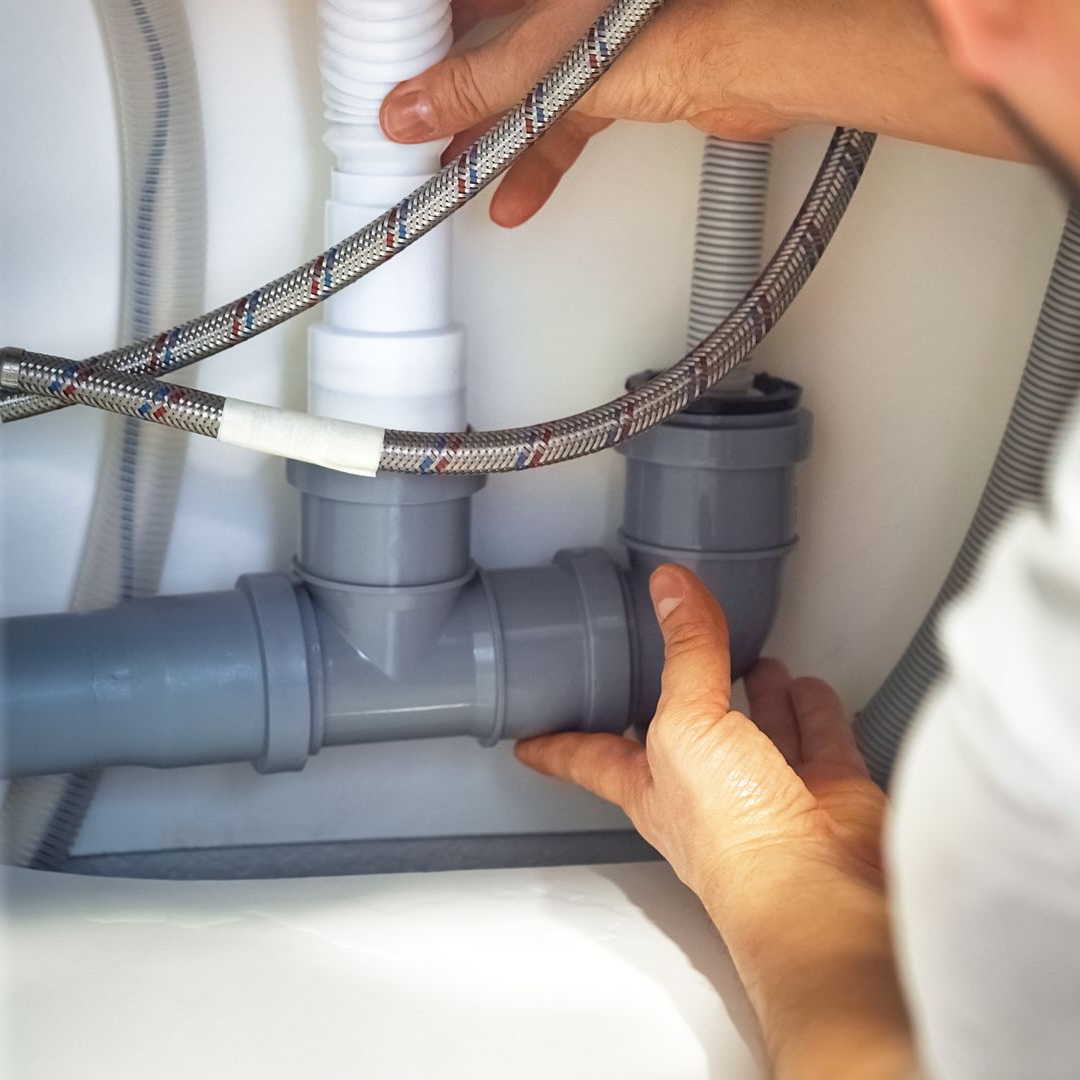 Des mains raccordent des tuyaux gris sous un évier blanc.