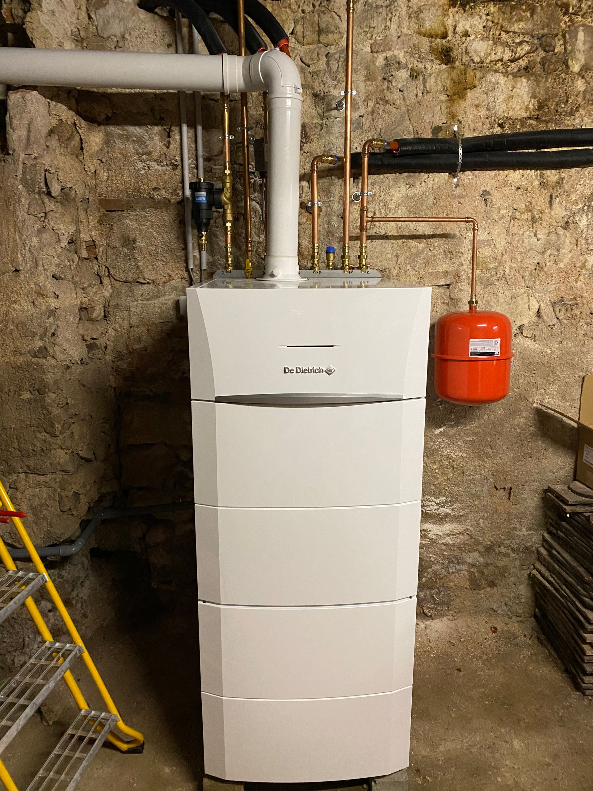 Chaudière blanche et vase d'expansion intégré au sous-sol. Les tuyaux sont raccordés à l'appareil contre un mur en pierre.