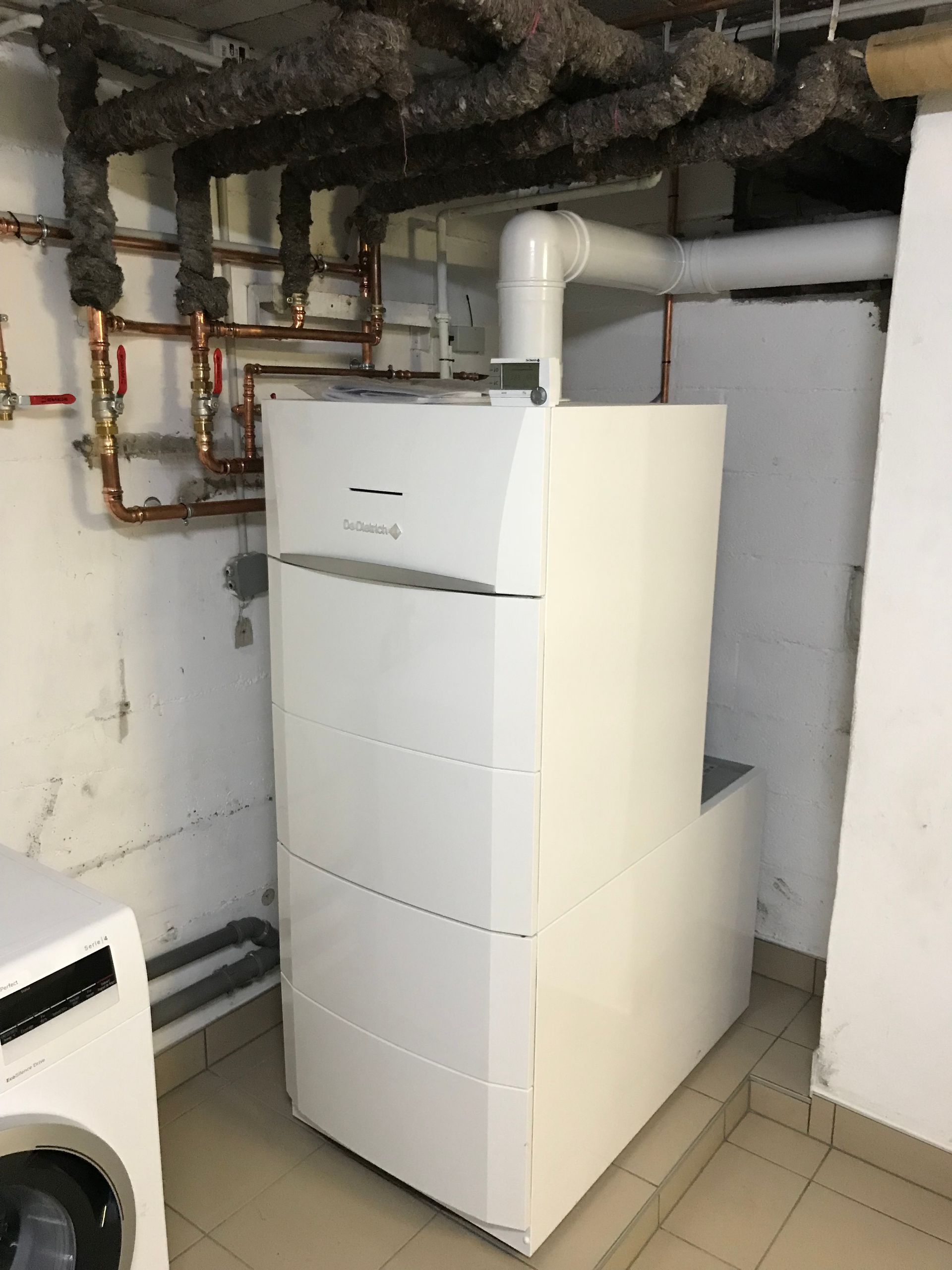 Système de chauffage blanc et ballon d'eau chaude contre un mur blanc, à côté d'une porte noire.