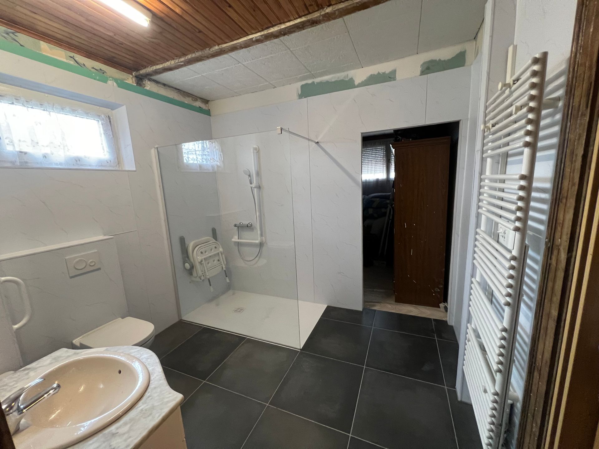 Rénovation de salle de bains : douche avec siège, toilettes, lavabo, sol carrelé, murs blancs et radiateur.